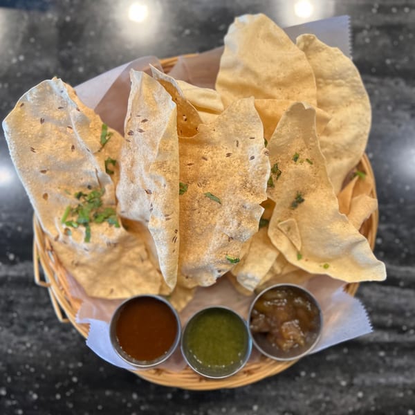 A basket of crispy papadum with three small bowls of dipping sauces on a dark table.