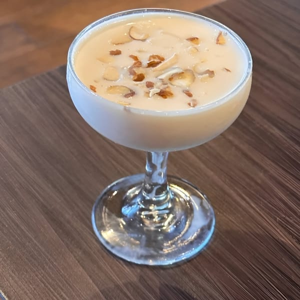 A creamy Indian dessert, possibly kheer, topped with sliced almonds in a stemmed glass on a wooden table.