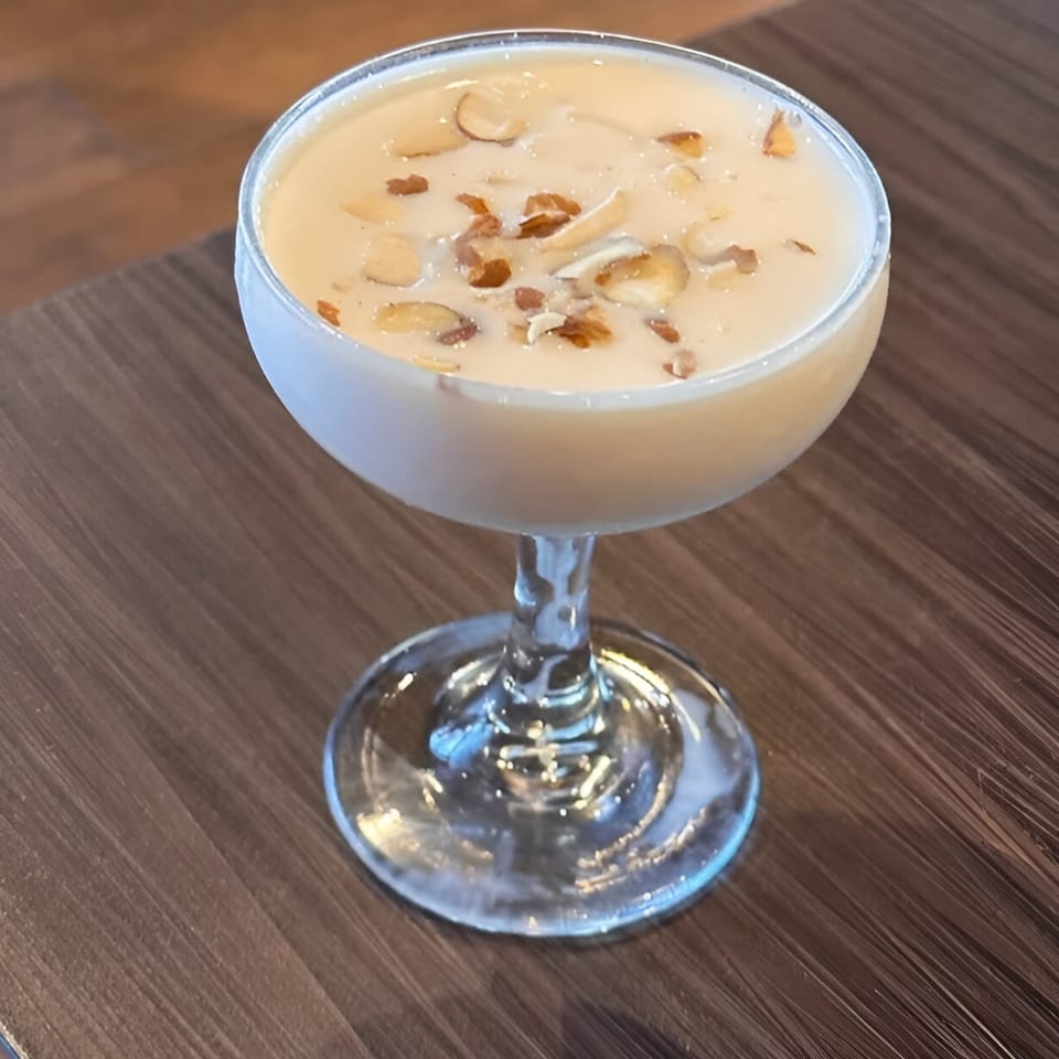 A creamy Indian dessert, possibly kheer, topped with sliced almonds in a stemmed glass on a wooden table.