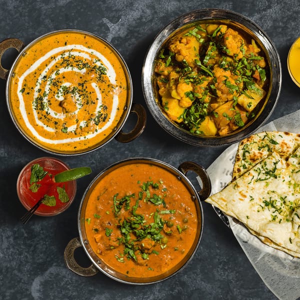 Overhead view of three Indian curries, naan bread, and a red cocktail on a dark surface.