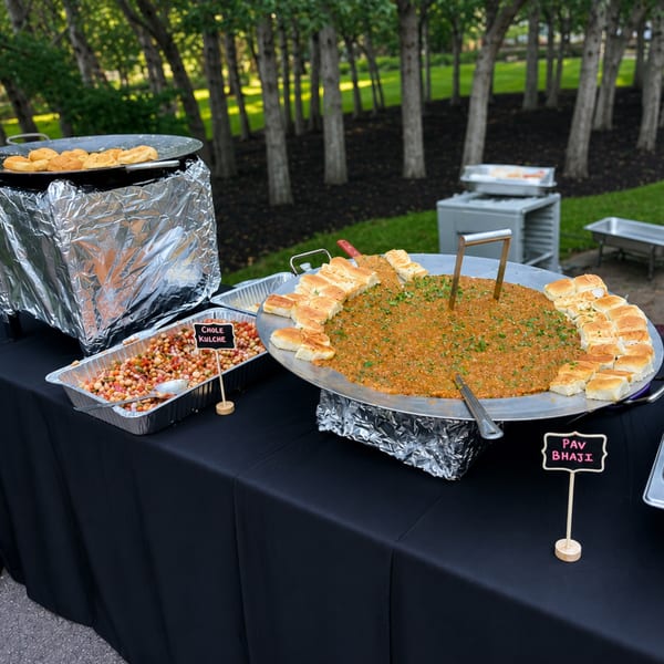 Outdoor catering setup with large pans of Pav Bhaji and Chole Kulche on a black-draped table.