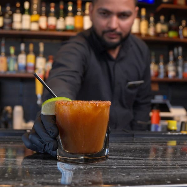 Bartender in black gloves serving a michelada with a lime wedge and chili-salted rim.