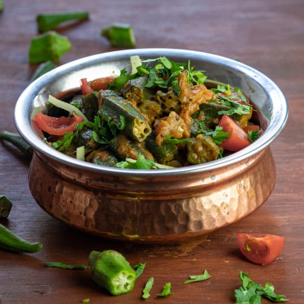 Okra curry with tomatoes and cilantro in a hammered copper bowl on a wooden surface.